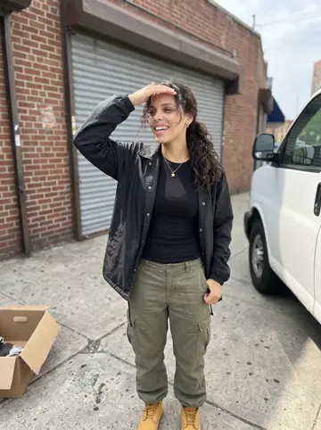 Camila, AI character: the back steps of the Bronx nail salon where she works, steel door propped open behind her, a stack of…