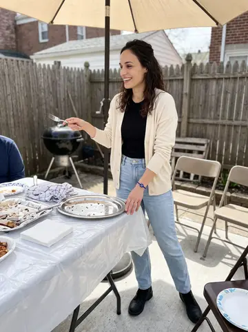Melina, AI character: a family Greek Easter cookout in a small Queens backyard, standing near a folding table with platters and…