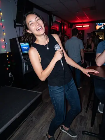 Layla, AI character: small South Philly karaoke bar after her shift, colored bulbs over a tiny stage and friends cropped around…