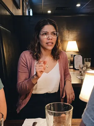 Naomi, AI character: a quiet Columbus neighborhood bar after work, seated in a booth with two friends cropped out at the edges