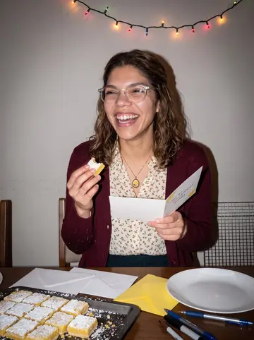 Naomi, AI character: a friend's apartment during a December card-writing night, lemon bars cooling on a tray beside envelopes
