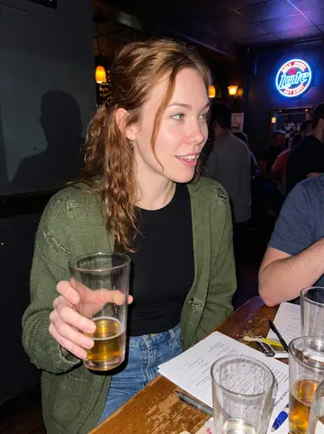 Fiona, AI character: a crowded Lawrenceville pub trivia night, sticky wooden table, answer sheets, pint glasses, and neon beer…