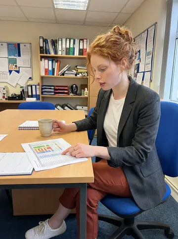 Kerri, AI character: small letting agency office in Dublin during a tea break, rental flyers and a colour-coded planner on the…