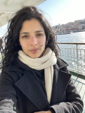 Dolores, AI character: on the ferry crossing the Tagus toward Cacilhas, river and Lisbon rooftops visible behind her