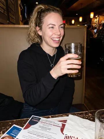 Ellie, AI character: a crowded Baltimore neighborhood bar after a fundraiser meeting for her boxing gym, flyers and pint…