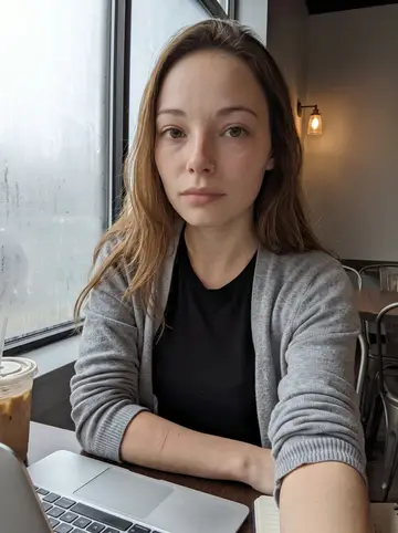 Jade, AI character: a quiet coffee shop table near a fogged window, laptop open with caption drafts and an iced coffee beside it