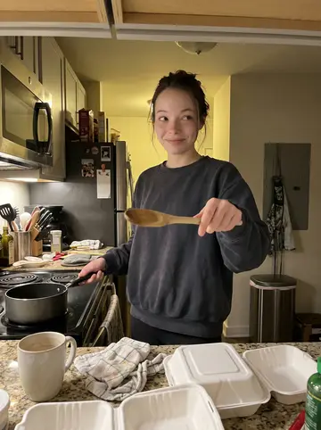 Jade, AI character: her small apartment kitchen in Nashville on a winter weeknight, takeout containers and a saucepan on the…