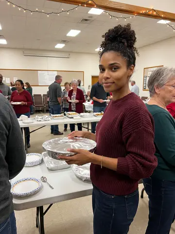 Jamila, AI character: a church hall holiday potluck in South Memphis, folding tables and warm string lights behind her