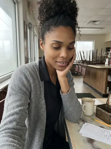 Jamila, AI character: a quiet corner table before opening, her tin of saved order slips and a half-finished coffee beside her