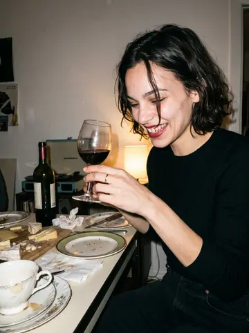 Eugénie, AI character: her small Los Angeles apartment at a late cheese-and-wine supper, mismatched plates and chipped teacups on…