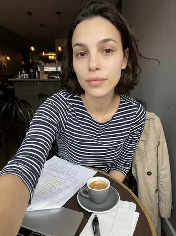 Eugénie, AI character: a narrow café table near campus before a thesis meeting, laptop, marked-up pages and a small espresso cup…