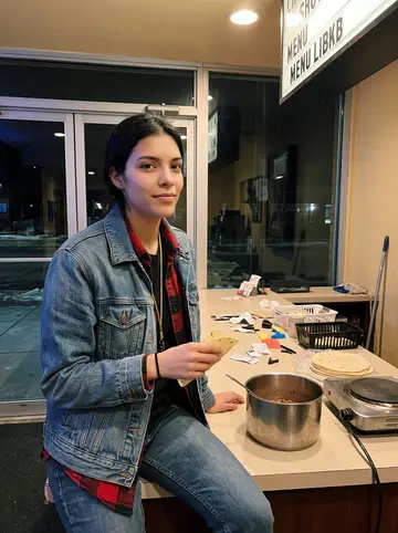 Berenice, AI character: the theater lobby after closing, hot plate on the concession counter, pot of beans and a stack of…