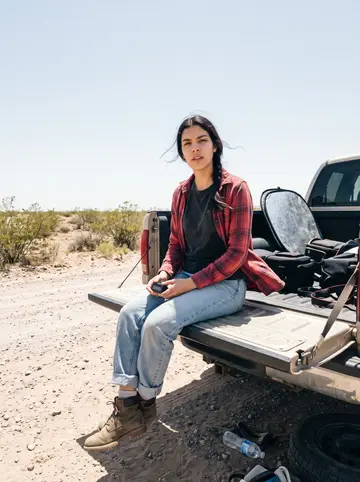 Berenice, AI character: a dusty roadside outside El Paso near low desert scrub, camera cases and a folded reflector in the back of…
