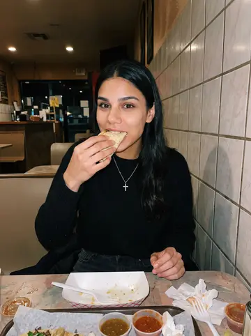 Yoselin, AI character: small late-night taco spot near her apartment, plastic table, salsa cups, bright wall tile behind her