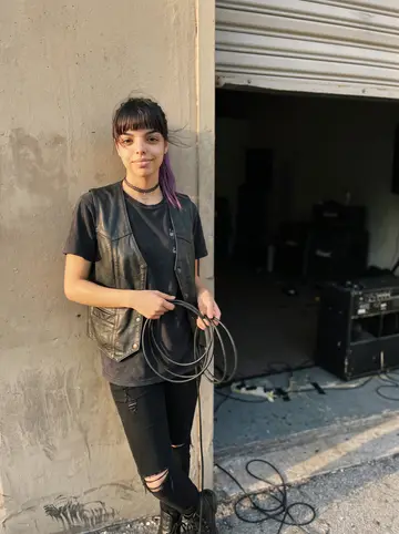Mayelin, AI character: outside a small Miami warehouse rehearsal space before a weekend show, the roll-up door half open behind her