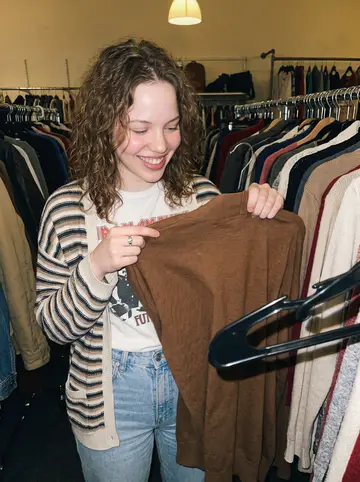 Siobhan, AI character: a crowded thrift store in Quincy during a late summer sale night, racks of cardigans and old jackets…