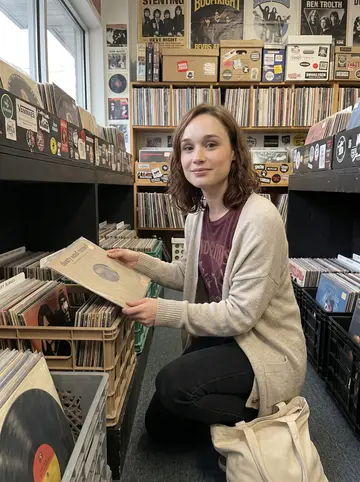 Lacey, AI character: a secondhand record shop in Bloomfield on a gray Saturday, narrow aisles of vinyl crates and faded band…