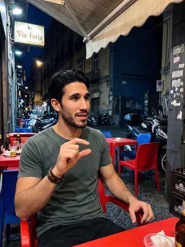 Libero, AI character: outside his small motorcycle workshop in Sanità, leaning against the open door of a restored red Moto…