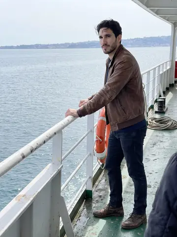 Libero, AI character: on a small ferry to Procida, standing by the rail with the Bay of Naples and pale spring water behind him