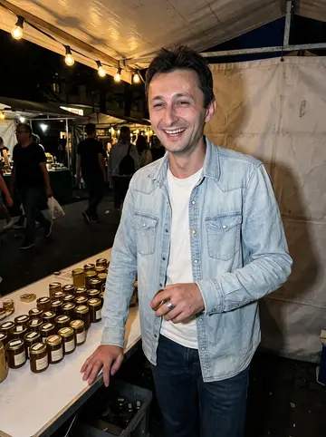 Hryhoriy, AI character: evening street market stall in Neukölln, small honey jars on a folding table, people passing behind him