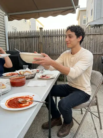 Enrico, AI character: his nonna's small backyard in East Boston during Sunday dinner, folding table crowded with plates and…