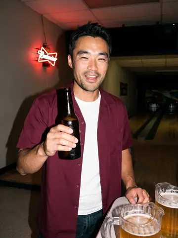 Caleb, AI character: a lanes bar in Bay View on league night, beer pitchers on the table and bowling lanes blurred behind him