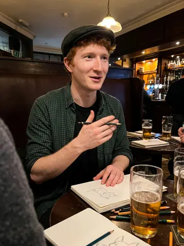Michel, AI character: a snug Glasgow pub during his monthly drawing night, small round table covered with sketchbooks and pint…