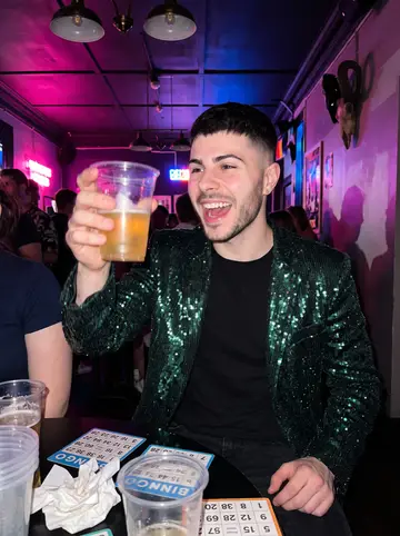Karol, AI character: inside a busy Church Street bar after drag bingo, colored lights, bingo cards and plastic cups on the table
