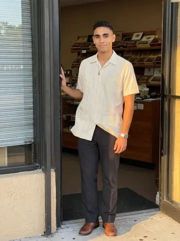 Osmany, AI character: outside his family's cigar shop in Hialeah at closing time, standing half in the doorway with the metal…