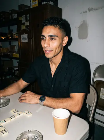 Osmany, AI character: the back room of the shop during a late dominoes game, plastic table crowded with tiles, coffee cups and…