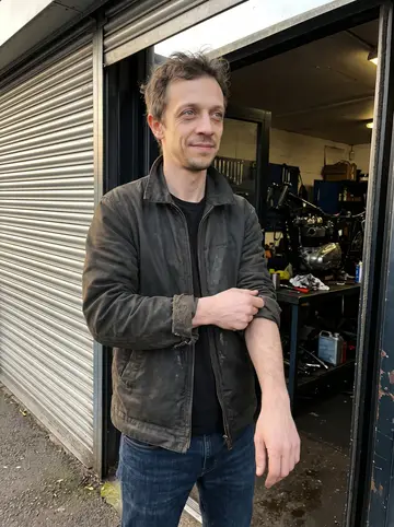 Dion, AI character: outside his Belfast motorcycle repair shop at closing time, the shutter half down and a rebuilt Triumph…