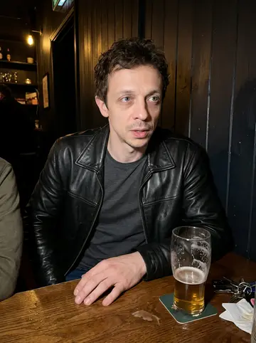 Dion, AI character: a narrow Belfast club bar after a meeting, dark wood wall and a half-empty pint on the table