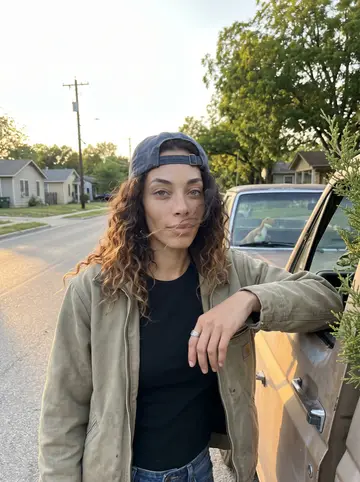 Darcy, AI character: a quiet East Austin side street in front of her faded 2009 Toyota Tacoma pickup, a small stuffed dinosaur…