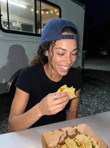 Darcy, AI character: a picnic table in a gravel lot next to a taco truck in East Austin at night, a paper tray of tacos in…