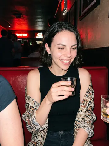 Victoria, AI character: a crowded Portland dive bar after a tiny gallery opening, standing near a red vinyl booth with friends…