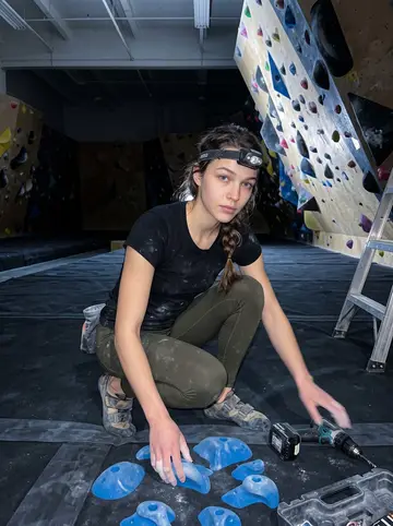 Kirsty, AI character: the empty climbing gym after closing, route-setting ladder, drill case, colored holds, and a headlamp beam…