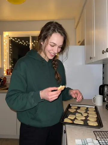 Kirsty, AI character: her small Toronto apartment kitchen at night, trays of uneven shortbread cooling on the counter and…