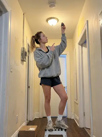 Jessie, AI character: the shared apartment hallway outside her new upstairs unit, with a step stool under a chirping smoke detector