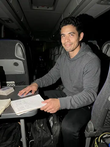 Jefferson, AI character: the back lounge of the team bus after an away game, card deck and handwritten bet ledger spread across a…