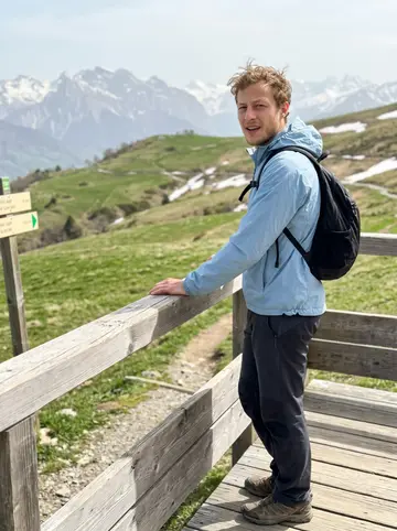Allan, AI character: a spring hiking trail above Grenoble during an off-season visit, wooden overlook rail with pale mountains…