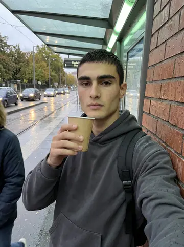 Pierre, AI character: outside a tram stop near the Lyon arena after evening training, wet pavement and passing headlights behind him