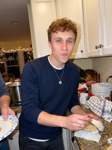 Colin, AI character: his family's crowded Gloucester kitchen during a noisy holiday meal, paper plates and a chowder pot on the…