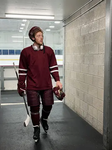 Gregor, AI character: back row of the team bus on an away trip, hockey bags piled in the aisle and grey highway visible through…