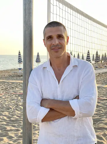 Fabio, AI character: the beach volleyball court of a family beach club in Rimini at sunset, leaning one shoulder on the post of…