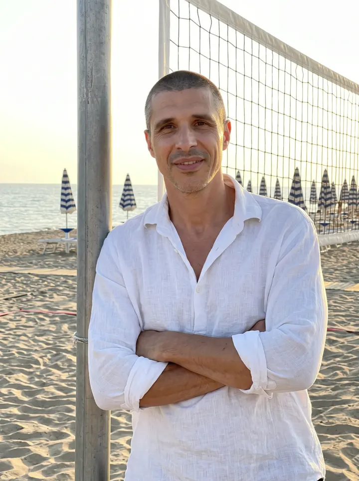 Fabio, AI character: the beach volleyball court of a family beach club in Rimini at sunset, leaning one shoulder on the post of…