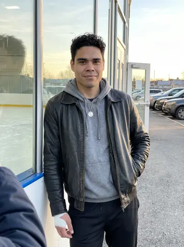 Cristóbal, AI character: outside the rink after morning skate, leaning against the sunlit glass by the player entrance with the…
