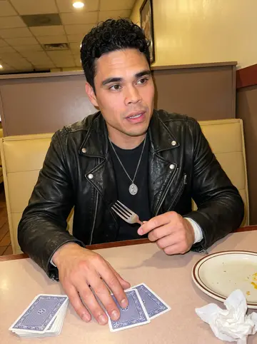 Cristóbal, AI character: a late-night diner booth after an away game, vinyl seats, half-finished plates, and a deck of cards on the…