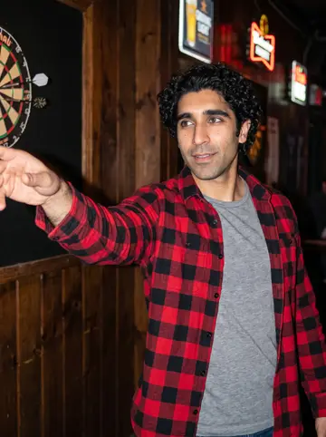 Kian, AI character: the dartboard corner of a Boston neighborhood bar called the Stone Well, wood paneling and beer signs behind