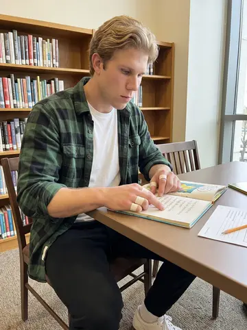 Brad, AI character: a public library tutoring table on Tuesday evening, picture books and worksheet pages spread across the…