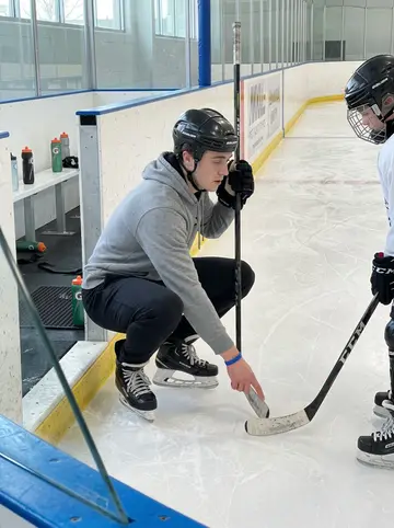 Ronald, AI character: Tuesday youth hockey clinic at a local rink, near the bench with rink glass and skate marks behind him